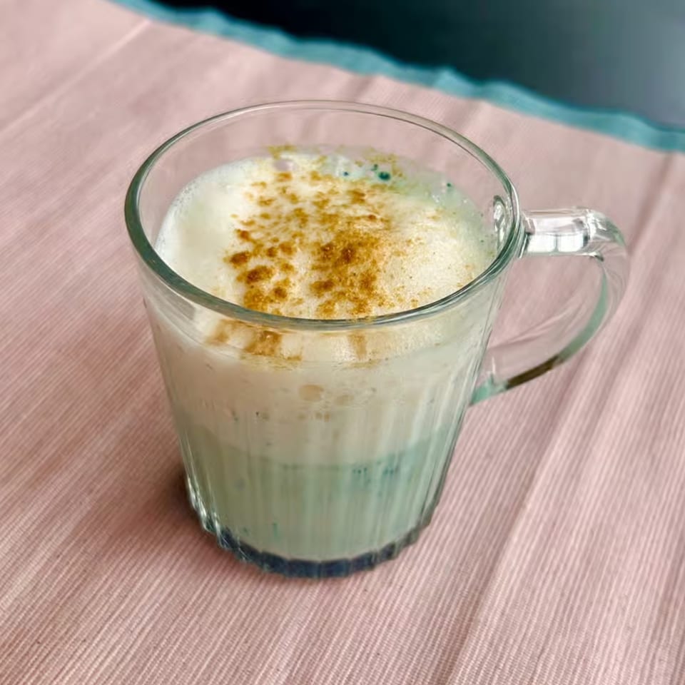 A clear glass mug filled with a light blue drink topped with white foam and cinnamon.