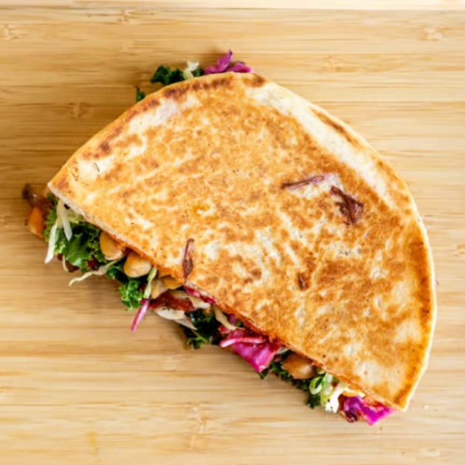 A golden-brown quesadilla, filled with green kale, red cabbage, and chickpeas, on a wooden surface.