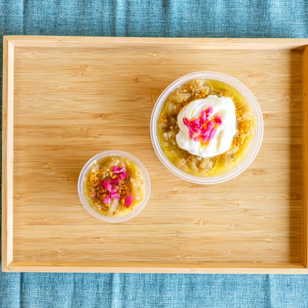 Two containers of Indian eggplant curry, one topped with yogurt and pickled onions, on a wooden tray.