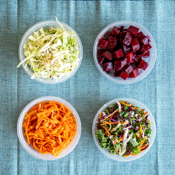 Four clear containers of fresh Indian side dishes: cabbage, beets, carrots, and mixed greens.