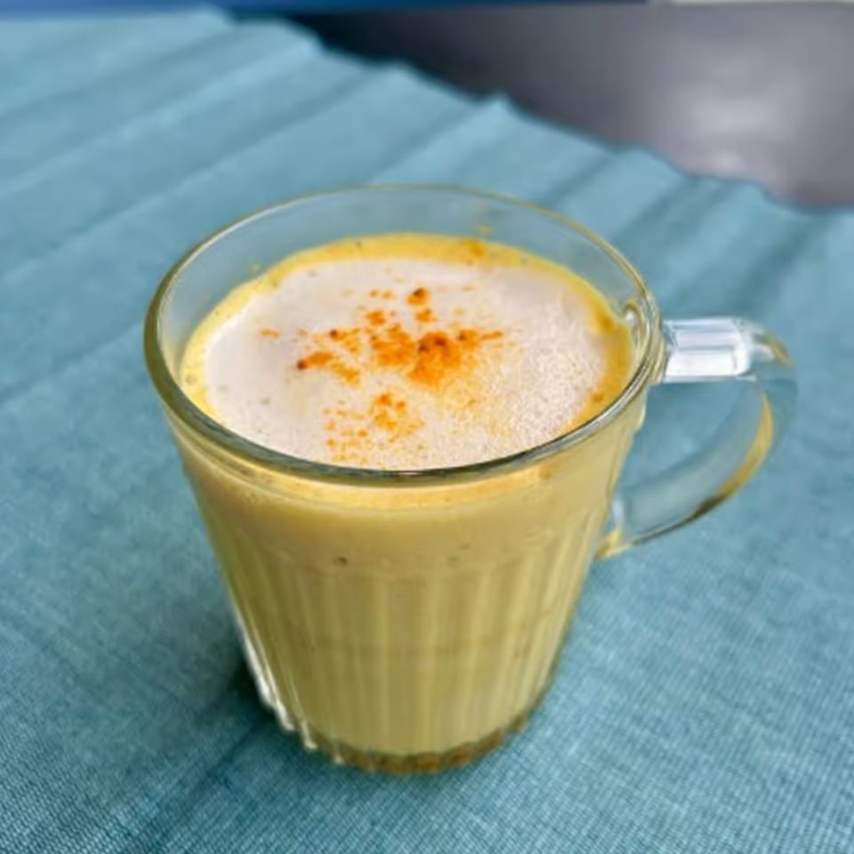 Golden milk latte with frothy top and spice sprinkles in a clear mug on a blue tablecloth.