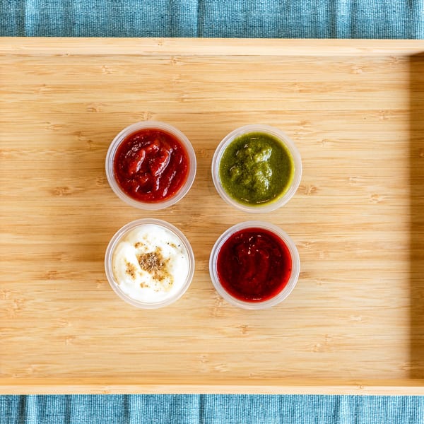 Four small plastic containers of Indian sauces, including red, green, and white, on a wooden tray.